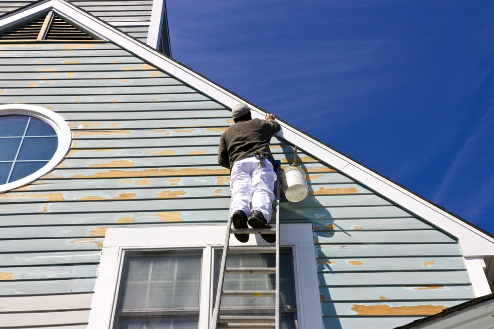 A technician sands worn and peeling areas of the home’s exterior siding while positioned on a ladder, preparing the surface for repainting and final repairs – Siding Repair in Indianapolis.