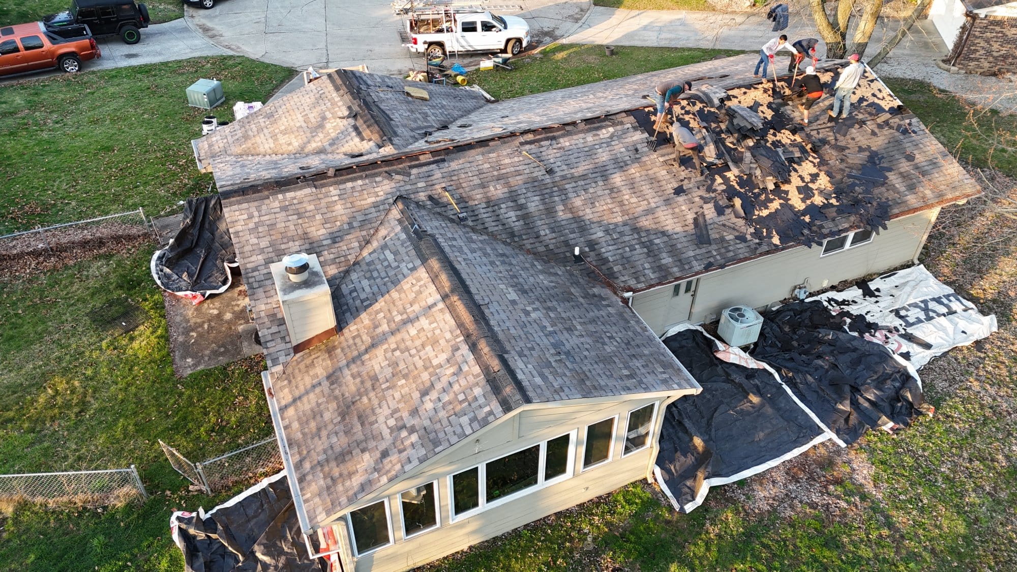 An aerial view of a house under roof renovation. Workers are removing old shingles, with tarps laid around the property to protect the ground and collect debris. Equipment and trucks are visible nearby - Copes R&R Construction An aerial view of a house under roof renovation. Workers are removing old shingles, with tarps laid around the property to protect the ground and collect debris. Equipment and trucks are visible nearby - Copes R&R Construction