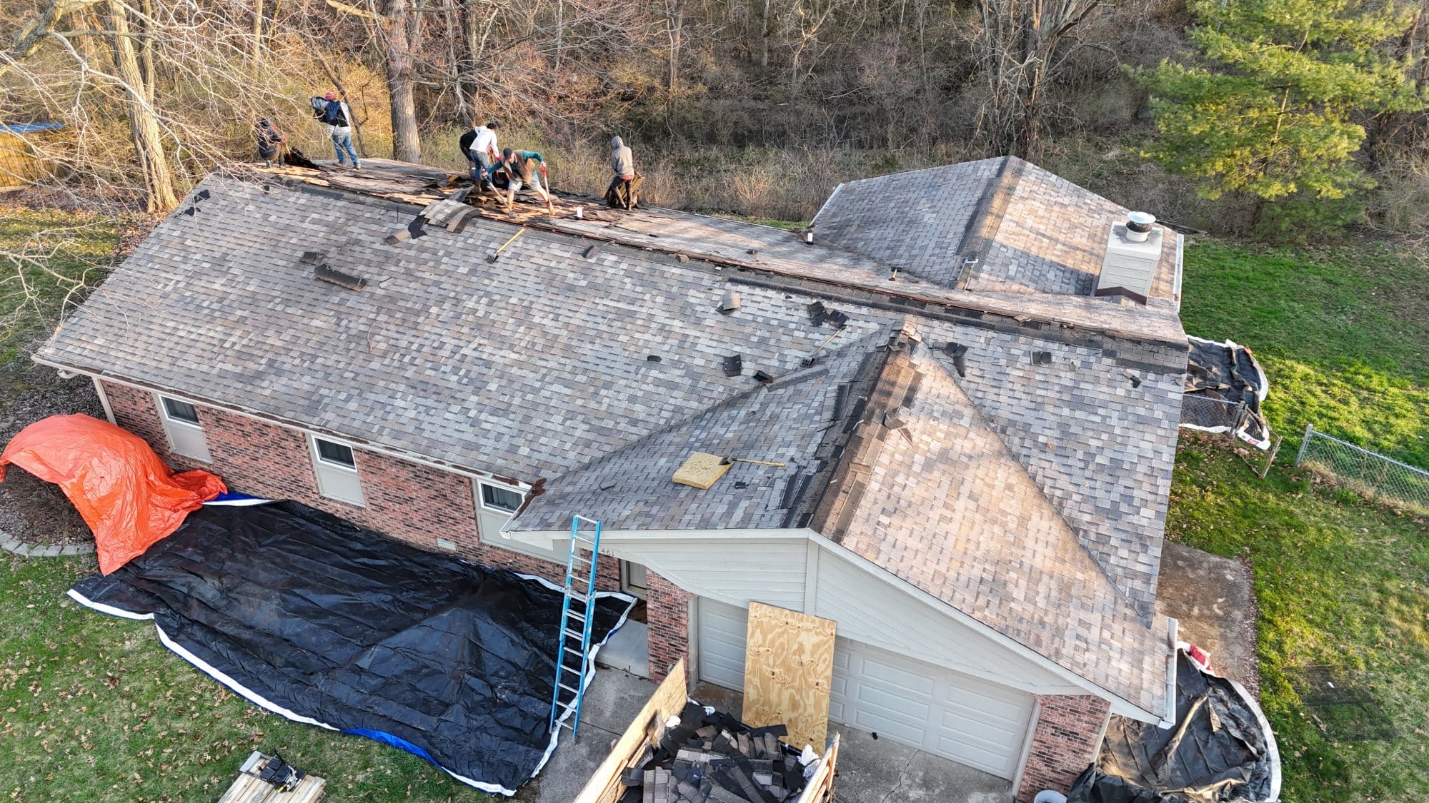 Aerial view of a house with roof repair in progress, showing workers removing shingles and protective tarps covering the surrounding area - Copes R&R Construction Aerial view of a house with roof repair in progress, showing workers removing shingles and protective tarps covering the surrounding area - Copes R&R Construction