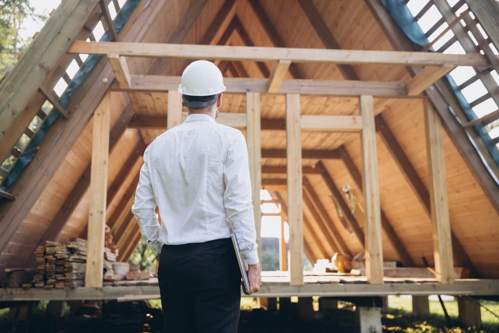 A construction professional wearing a hard hat evaluates the framing of an A-frame style house. The exposed wooden beams and open structure highlight an early stage of residential building development – General Housing Construction.