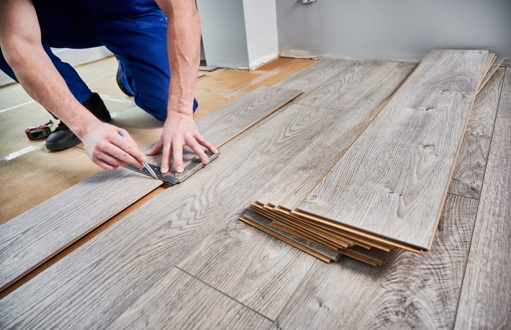 A close-up view of a contractor marking and cutting laminate planks on a subfloor, preparing each board for a precise fit. The gray wood-grain planks highlight a modern flooring upgrade in progress – Flooring.