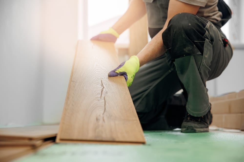 A flooring installer positions a long, wood-textured plank over an underlayment, preparing to lock it into place. The warm tone of the material creates a stylish and durable surface for the room – Flooring.