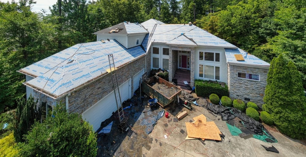 An overhead view of a large home undergoing roof replacement, with protective underlayment installed and roofing materials staged across the property. Ladders, tools, and debris outline an active professional roofing project. – Roof Replacement in Indianapolis