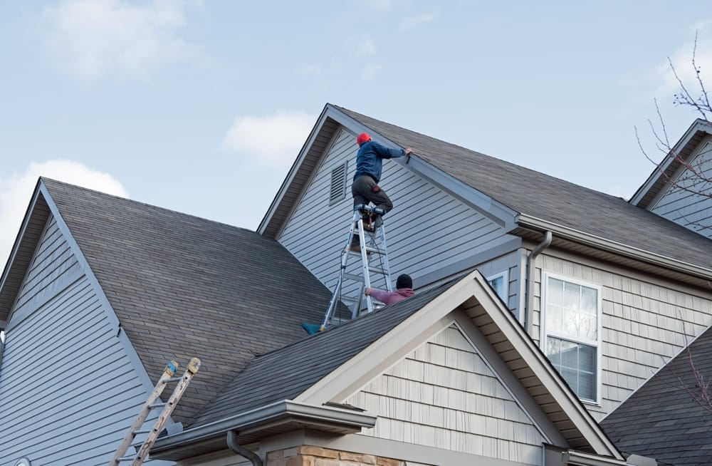 Two roofing contractors work on a tall residential roof using ladders and safety gear to perform repairs. The scene shows active maintenance on shingles and trim to restore the home’s exterior – Roof Repair in Indianapolis.