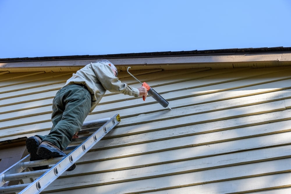 A contractor stands on a ladder applying caulk to damaged exterior siding, sealing gaps and preventing moisture intrusion to restore the home’s structural protection – Siding Repair in Indianapolis.