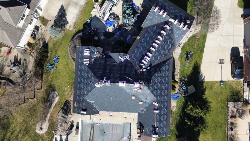 An overhead view of a house with roofing in progress. The roof is covered with underlayment and bundles of shingles are strategically placed across it. Surrounding the house are tools, equipment, and materials, with workers actively on-site - Copes R&R Construction An overhead view of a house with roofing in progress. The roof is covered with underlayment and bundles of shingles are strategically placed across it. Surrounding the house are tools, equipment, and materials, with workers actively on-site - Copes R&R Construction