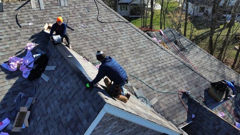 Workers on a roof installing shingles and securing materials, with tools and cables spread across the rooftop - Copes R&R Construction Workers on a roof installing shingles and securing materials, with tools and cables spread across the rooftop - Copes R&R Construction