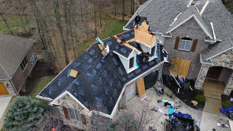 Aerial view of a house with roof repairs underway, showing underlayment, exposed wooden sections, and workers with tools on-site - Copes R&R Construction Aerial view of a house with roof repairs underway, showing underlayment, exposed wooden sections, and workers with tools on-site - Copes R&R Construction